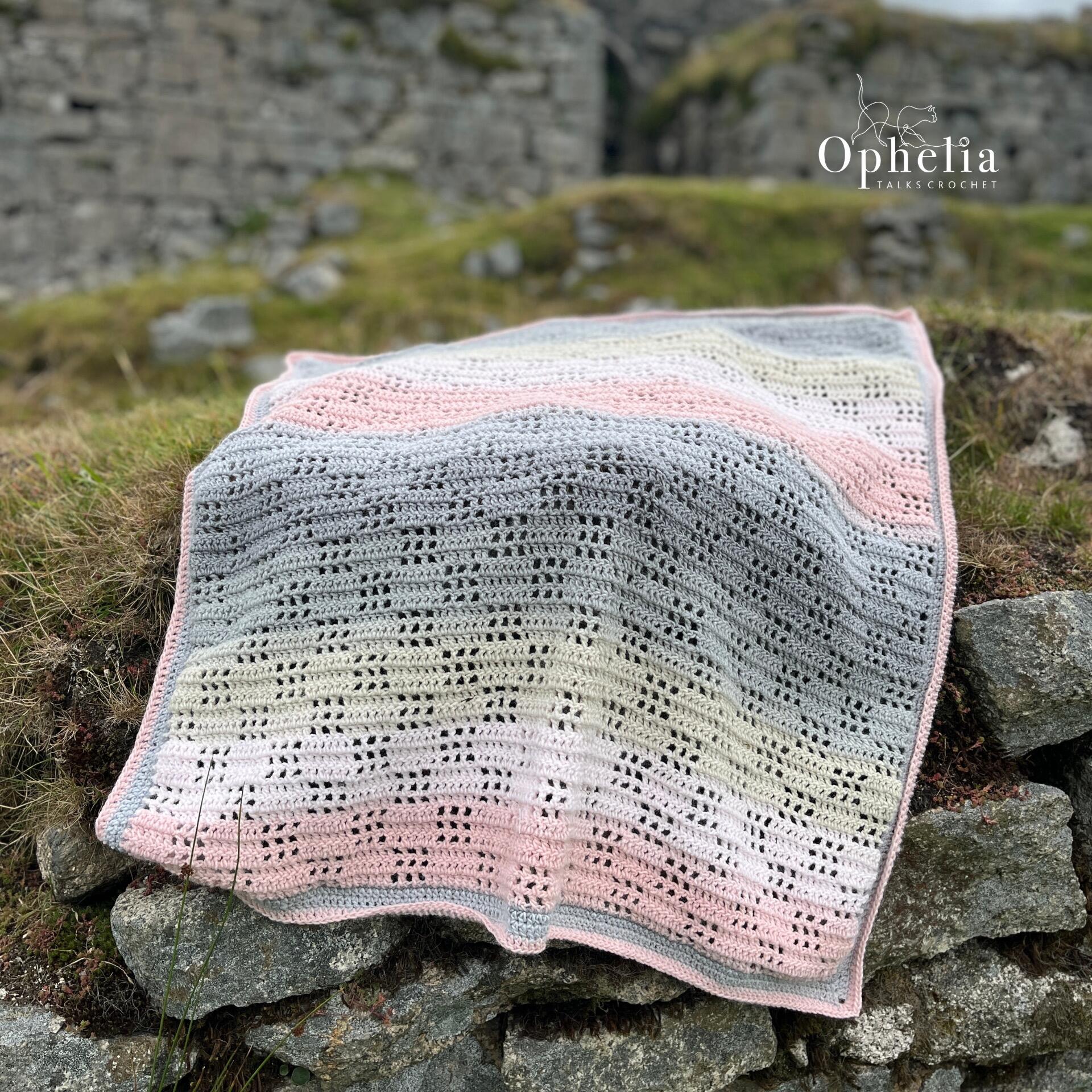 Handmade filet crochet baby blanket in soft pastel shades of pink, grey, and cream displayed on a stone wall outdoors. Created using the Ode To Baby crochet pattern by Ophelia Talks Crochet.