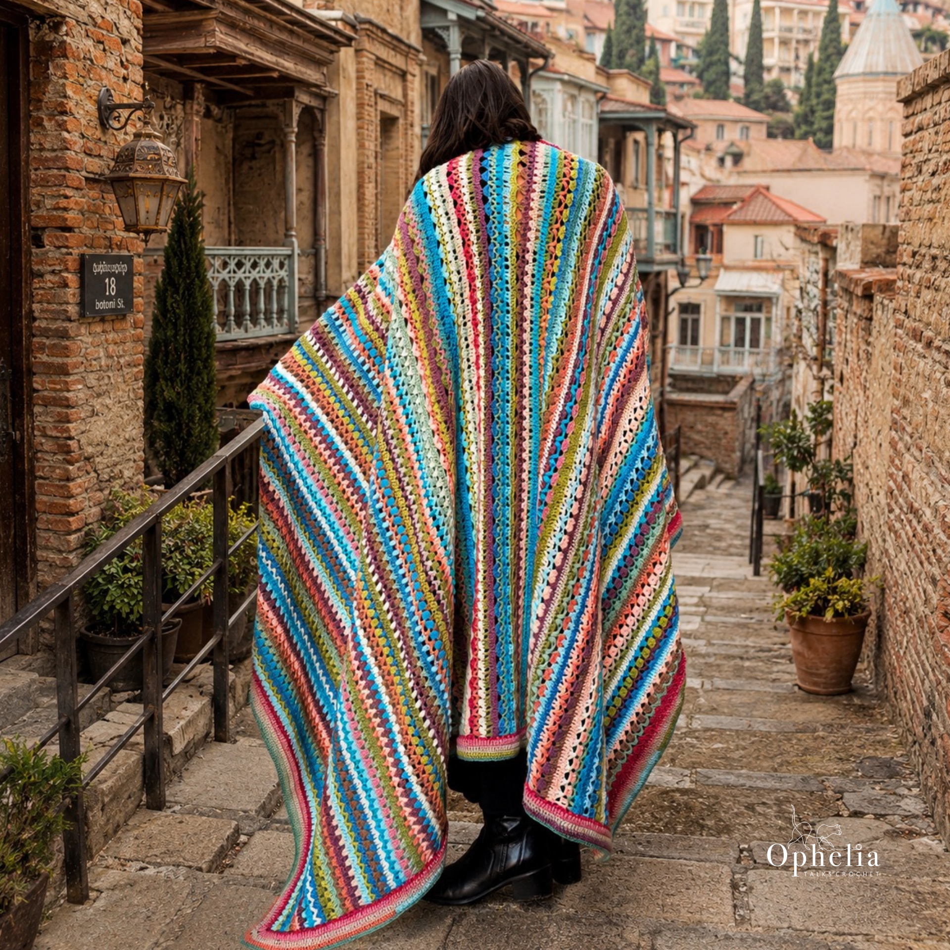 Woman seen from behind wearing a colourful striped crochet blanket wrap on a stone street in old Tbilisi, surrounded by rustic brick walls, traditional balconies and historic city architecture.