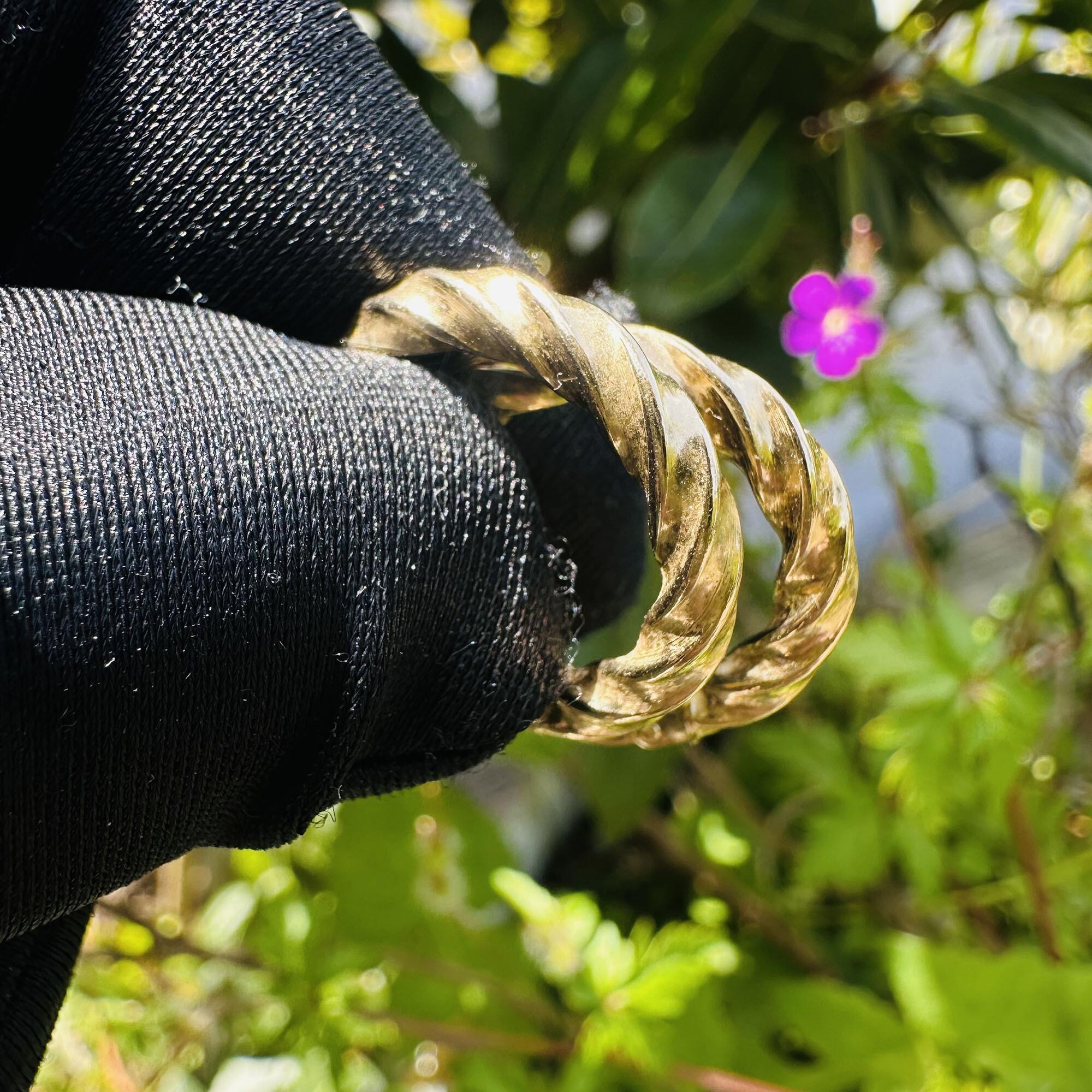 Italian 9k yellow gold twist rope hoop earrings held in black gloved fingers with garden background.