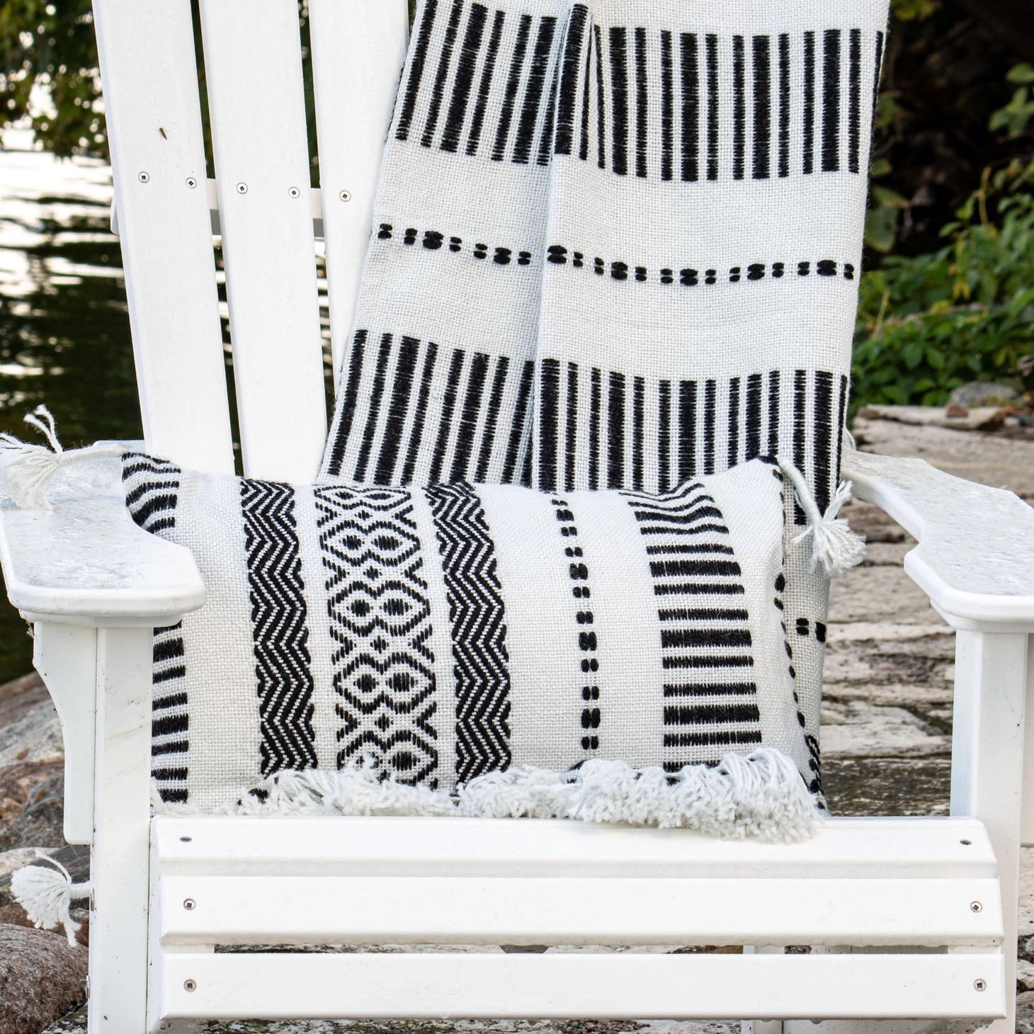 A hand woven outdoor boho cushion with a mix of black and white yarns and thread wrapped corner tassels, on a white chair.
