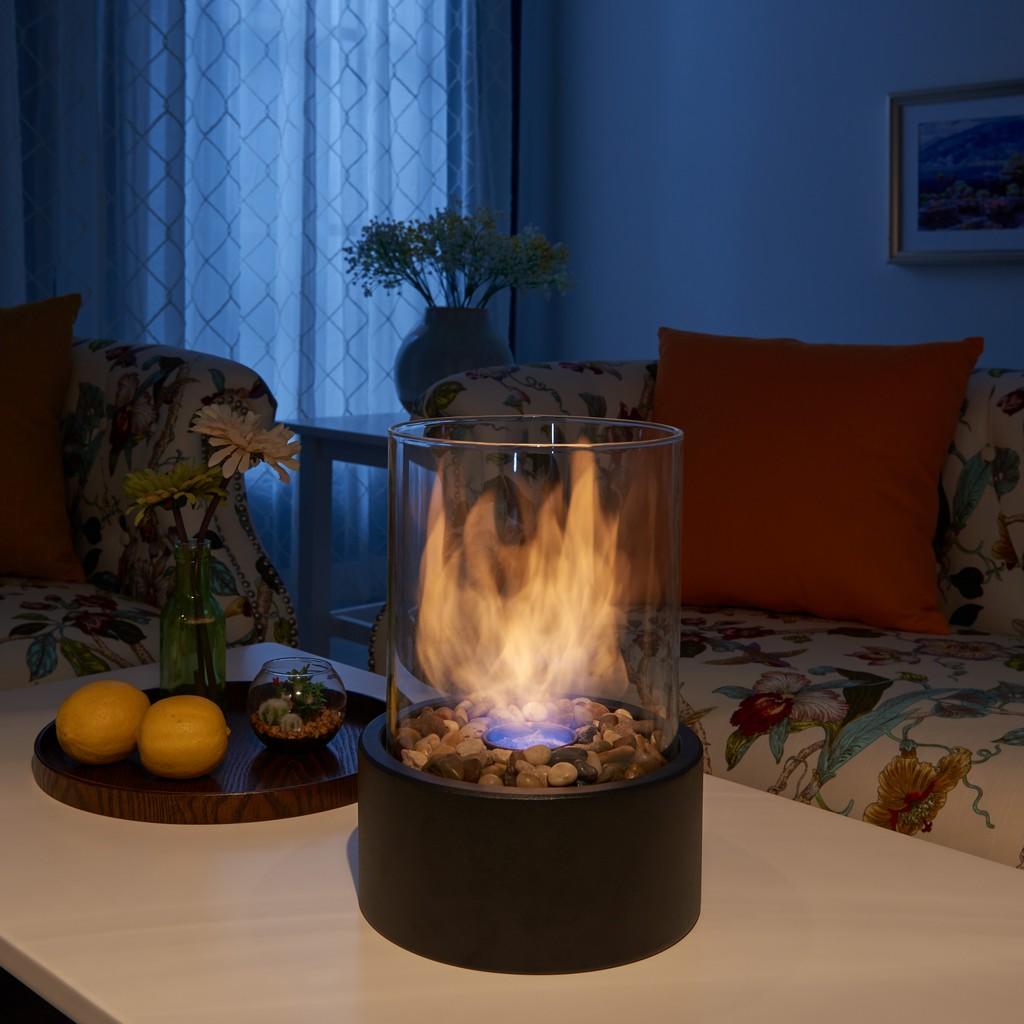 Lit portable tabletop fire pit with a black metal base, decorative rocks & a cylindrical glass body, on a living room table.