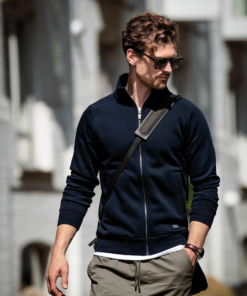 Man wears dark sunglasses, a dark zip-up jacket, and khaki pants while walking with a Nimbus N110M crossbody bag on a city street.