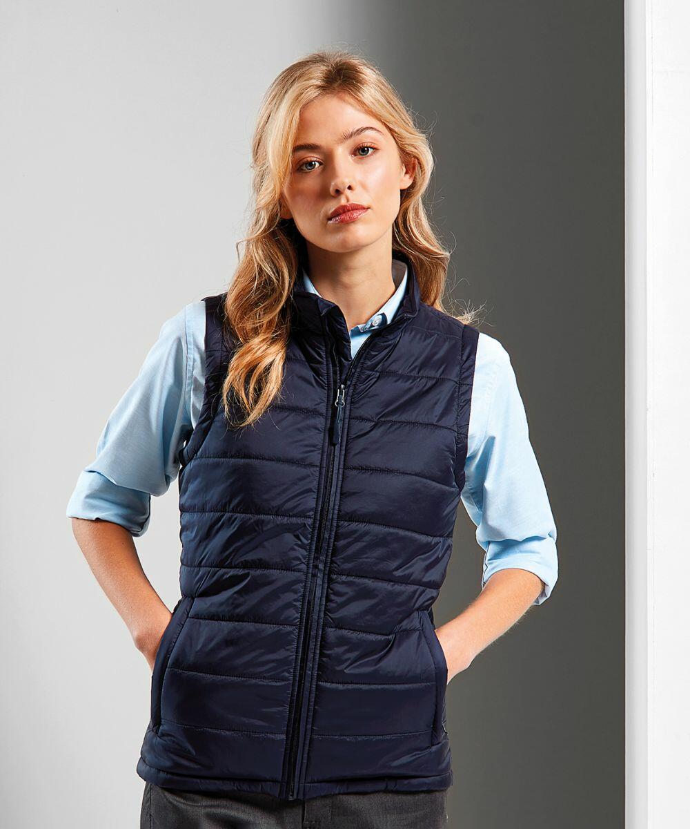A woman with blonde hair, wearing a light blue shirt and a dark blue Premier PR815 quilted vest, stands facing forward with hands in pockets.