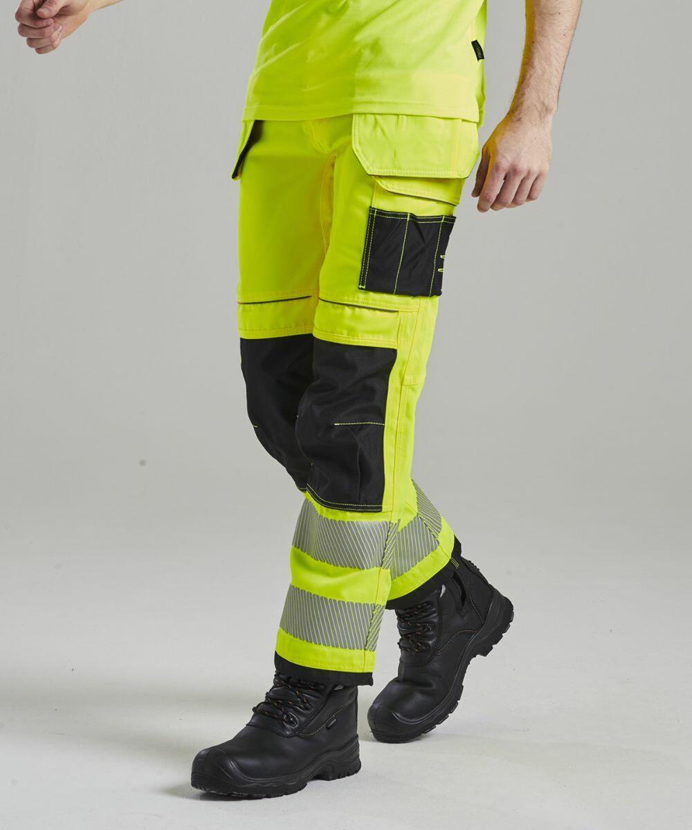 A person wearing Portwest PW367 high-visibility yellow work pants with black and silver reflective accents and black work boots.