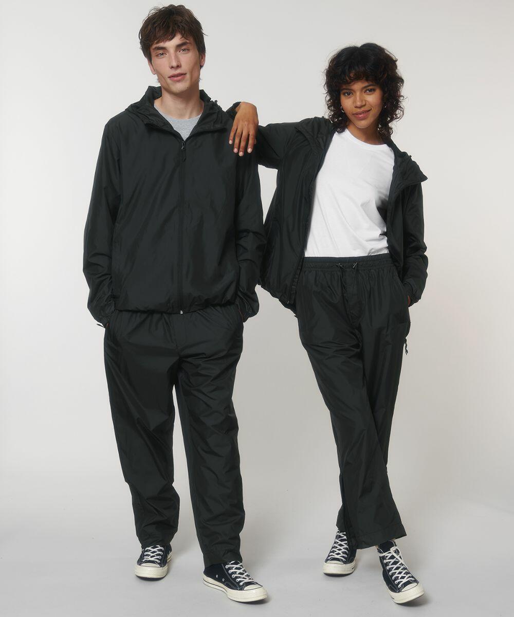 Two people are standing, both wearing black tracksuits and black sneakers with white laces. The person on the left has their arm around the person on the right, who is wearing a white Stanley/Stella SX171 shirt under the tracksuit jacket.