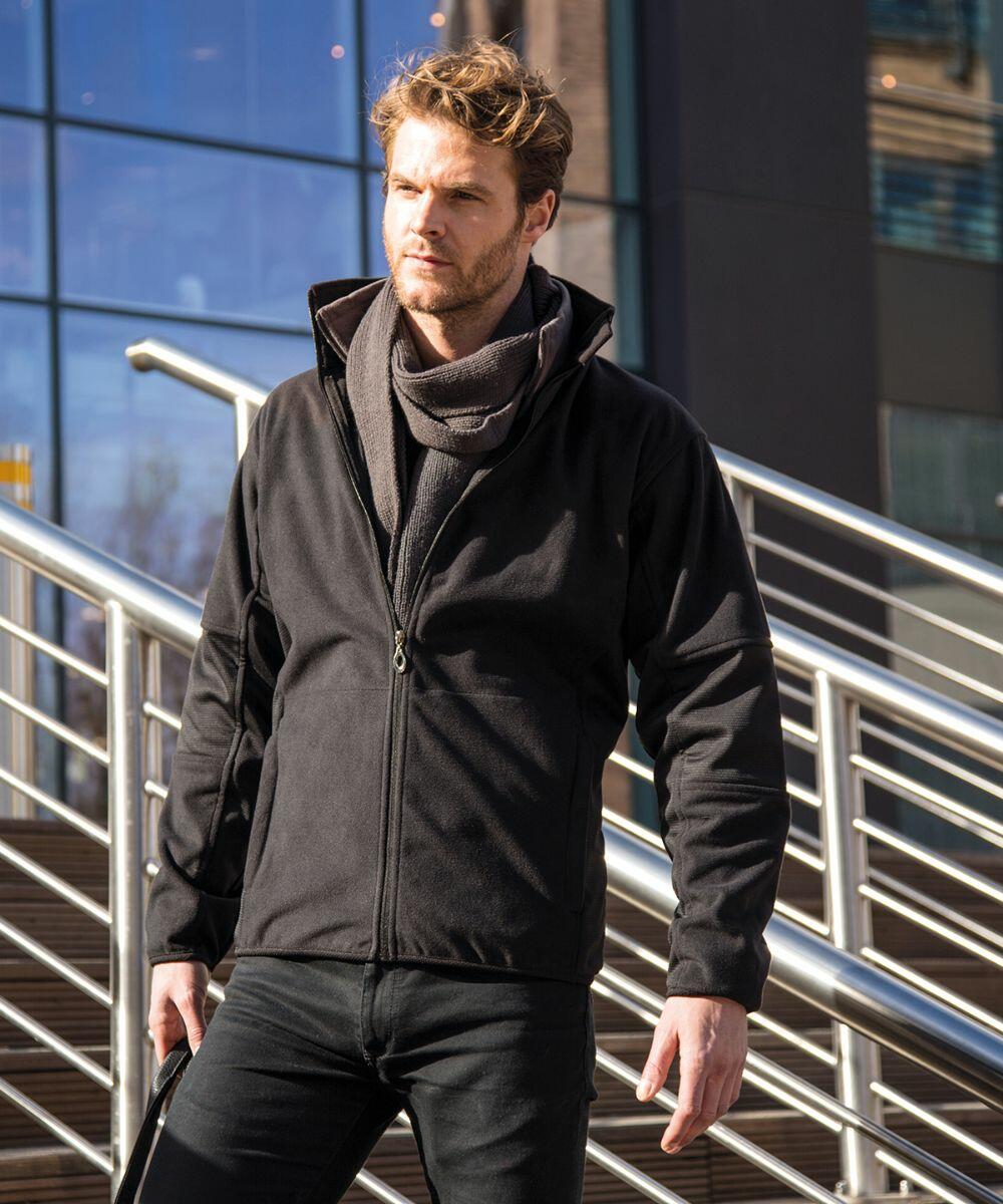 A man with a beard stands in front of a modern glass building, wearing a black jacket and scarf, holding a Result R131M bag. Metal railings and stairs are visible in the background.