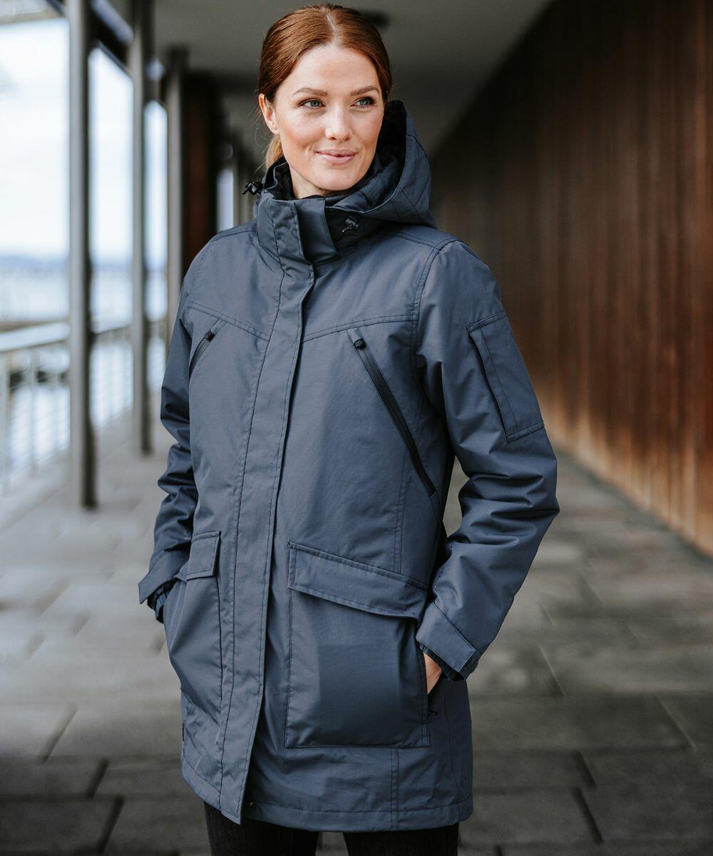 A person with brown hair stands outside, wearing a blue Stormtech ST217 insulated jacket with hands in pockets, looking to the side. The background features a walkway and wooden wall.