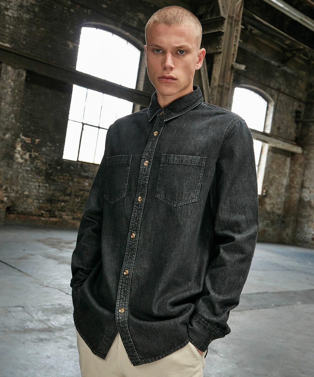 A person with short hair is standing indoors wearing a dark denim button-up BY152 shirt by Build Your Brand. The background features large windows and an industrial-style setting.