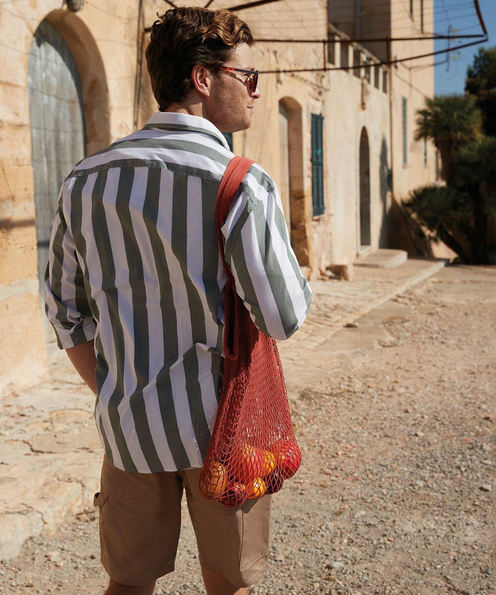 A man in a striped shirt and khaki shorts stands on a sunny street, carrying a red Westford Mill WM150 with oranges.