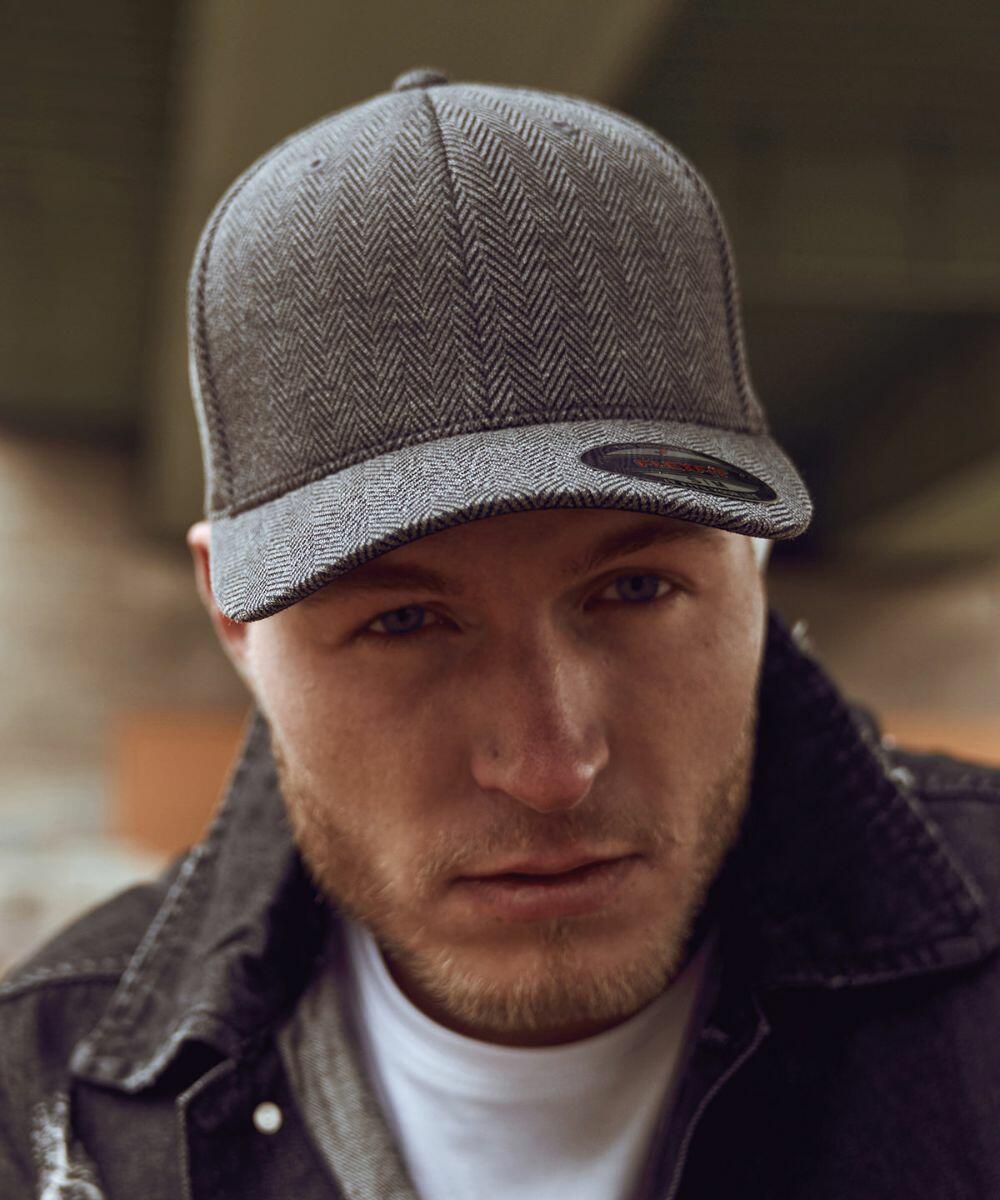 A man wearing a gray herringbone-patterned Flexfit By Yupoong YP108 baseball cap and a black denim jacket looks directly at the camera. Background is blurred.