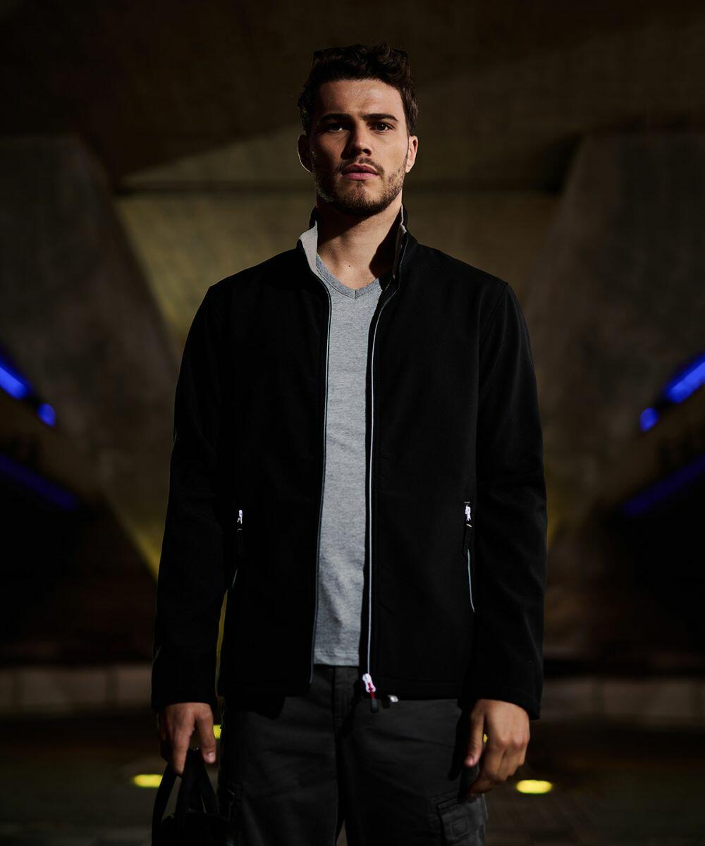 A man with short hair and a beard is standing under a bridge, wearing a light gray T-shirt, a black Regatta Professional RG590 jacket, and cargo pants. The background is dimly lit with blue and yellow lights.
