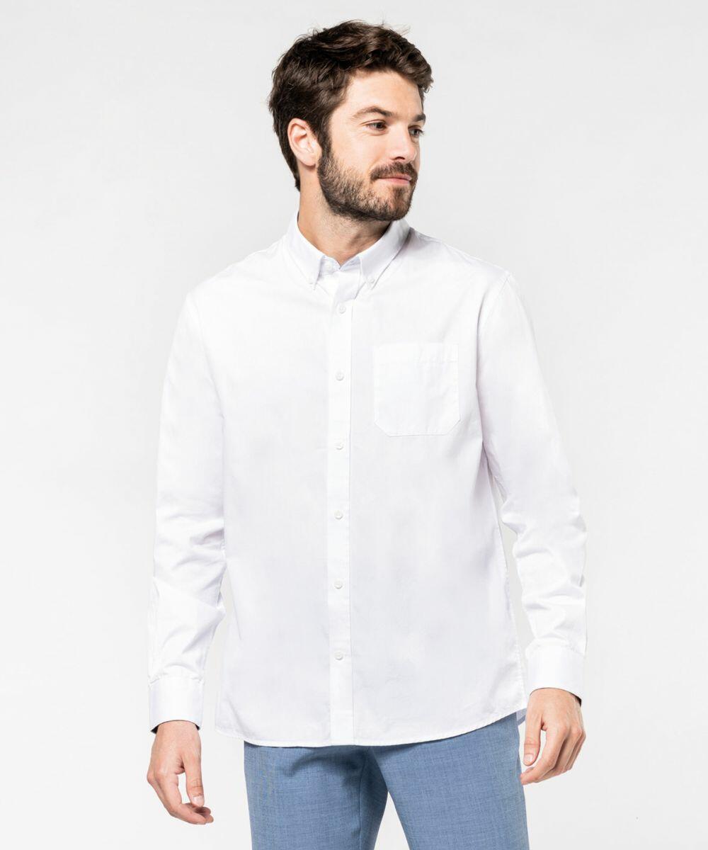 A man with short hair and a beard wears a white button-down KB586 by Kariban and light blue pants, standing against a plain white background, looking slightly to the side.