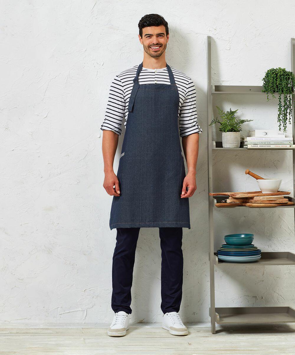 A person stands against a white wall, wearing a striped shirt, dark apron, dark pants, and white Premier PR122 shoes. Next to them is a shelf with dishes, books, and a plant.