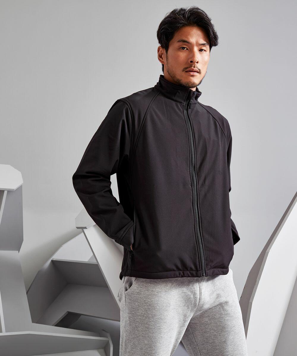 A man with short hair wearing a black zip-up 2786 TS012 jacket and light gray pants stands with hands in pockets, against a geometric background.
