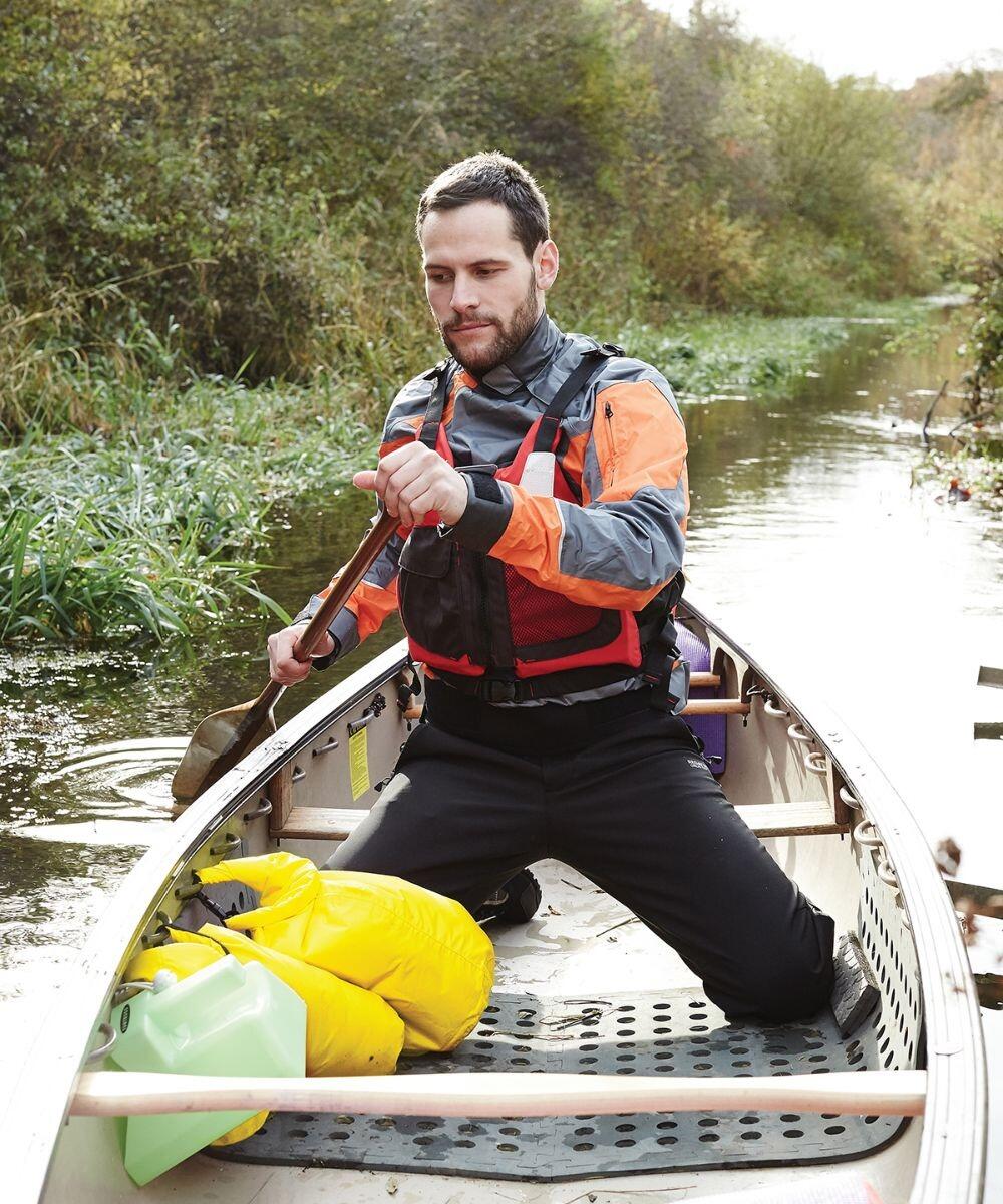 A person paddling a canoe in a narrow waterway, wearing a life jacket and surrounded by vegetation, with the Quadra QX615 dry bag and other gear in the canoe.