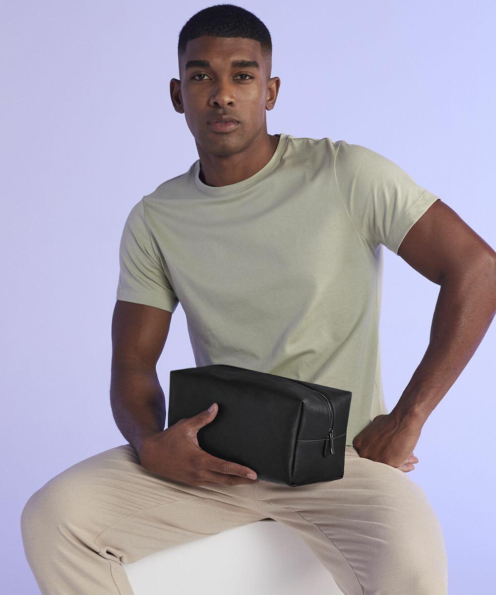 A person wearing a light olive green T-shirt and beige pants is sitting on a white surface against a light purple background. They are holding a black Bagbase BG749 zippered pouch in their lap with both hands. They are looking directly at the camera.
