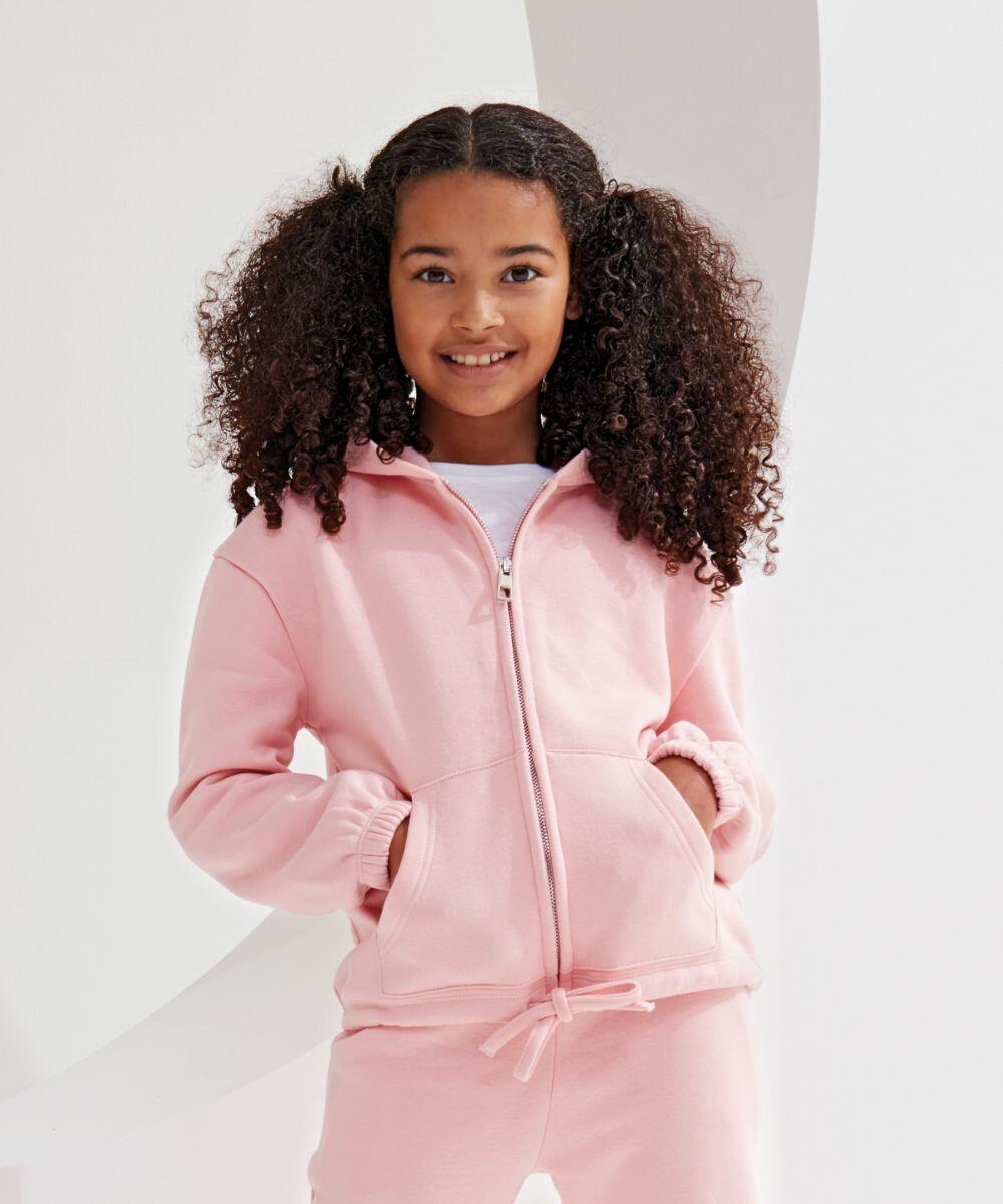 A young girl with curly hair stands smiling in a TriDri TR61B pink zip-up hoodie and matching pants with her hands in her pockets.