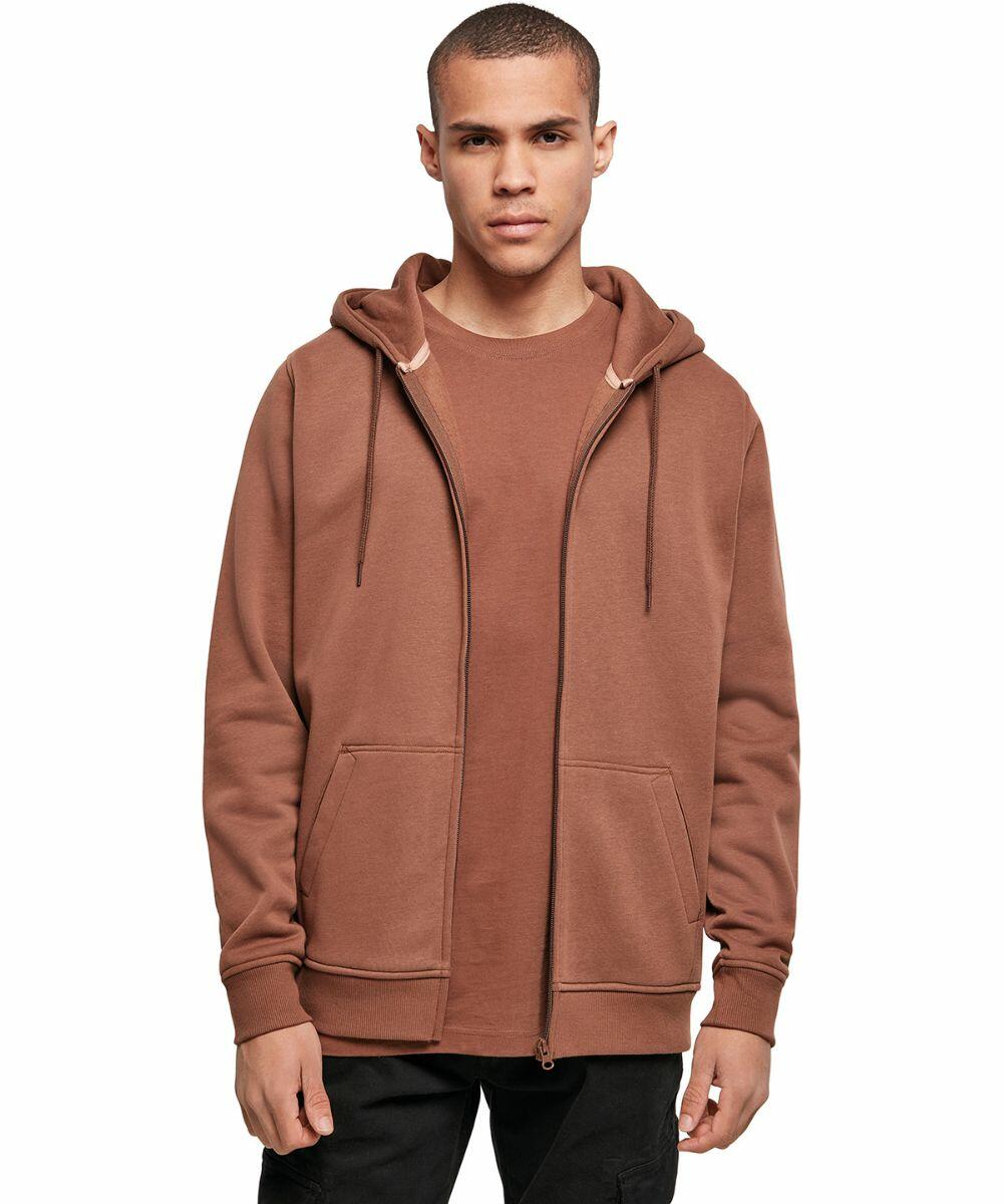 A person stands facing the camera, wearing a brown Build Your Brand BY012 zip-up hoodie over a matching brown t-shirt. The hoodie has drawstrings and two large front pockets. The person has short hair and a neutral expression. The background is plain and white.