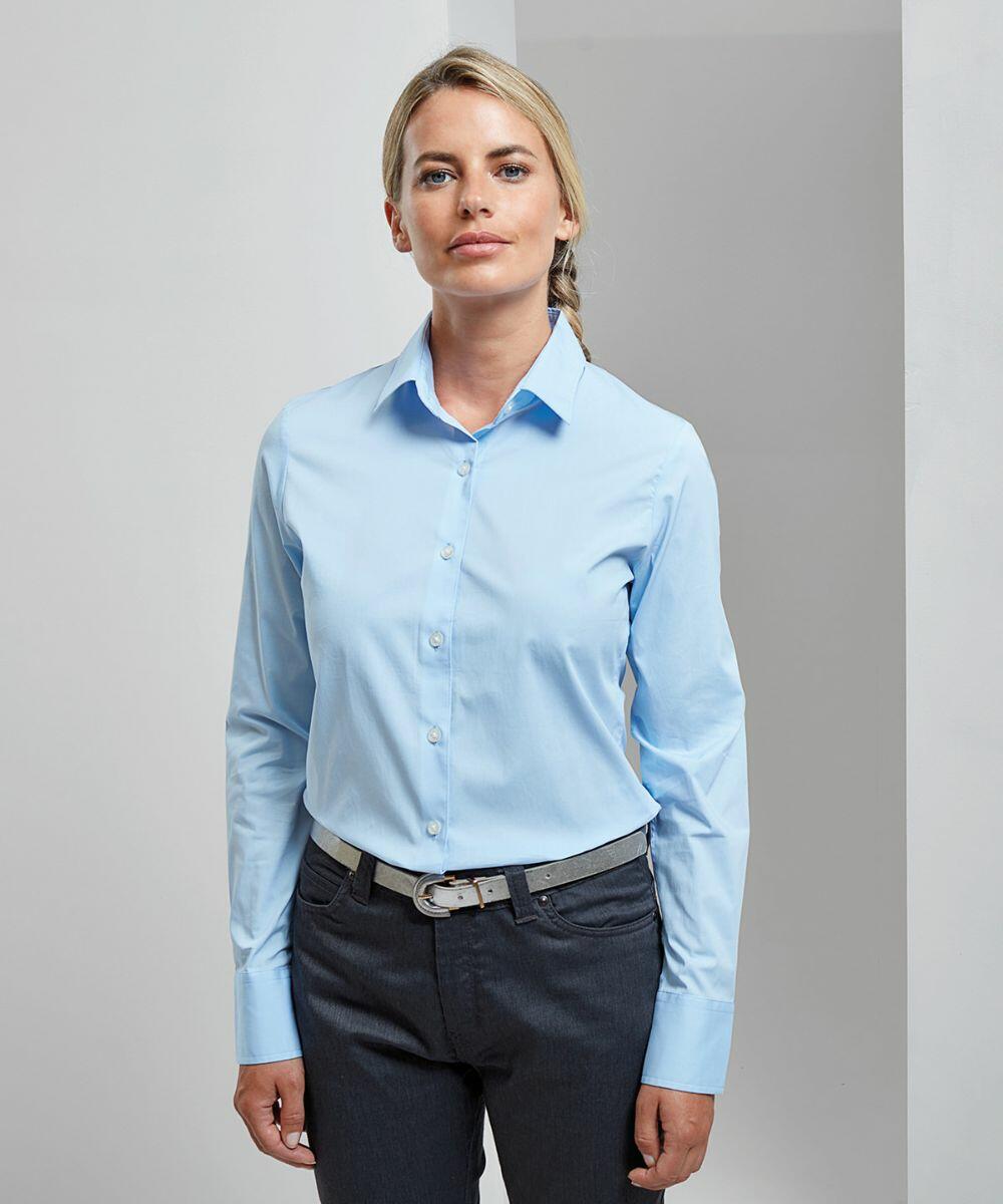 A person with blond hair wearing a light blue, long-sleeved Premier PR344 button-up shirt and dark gray pants stands against a plain background.