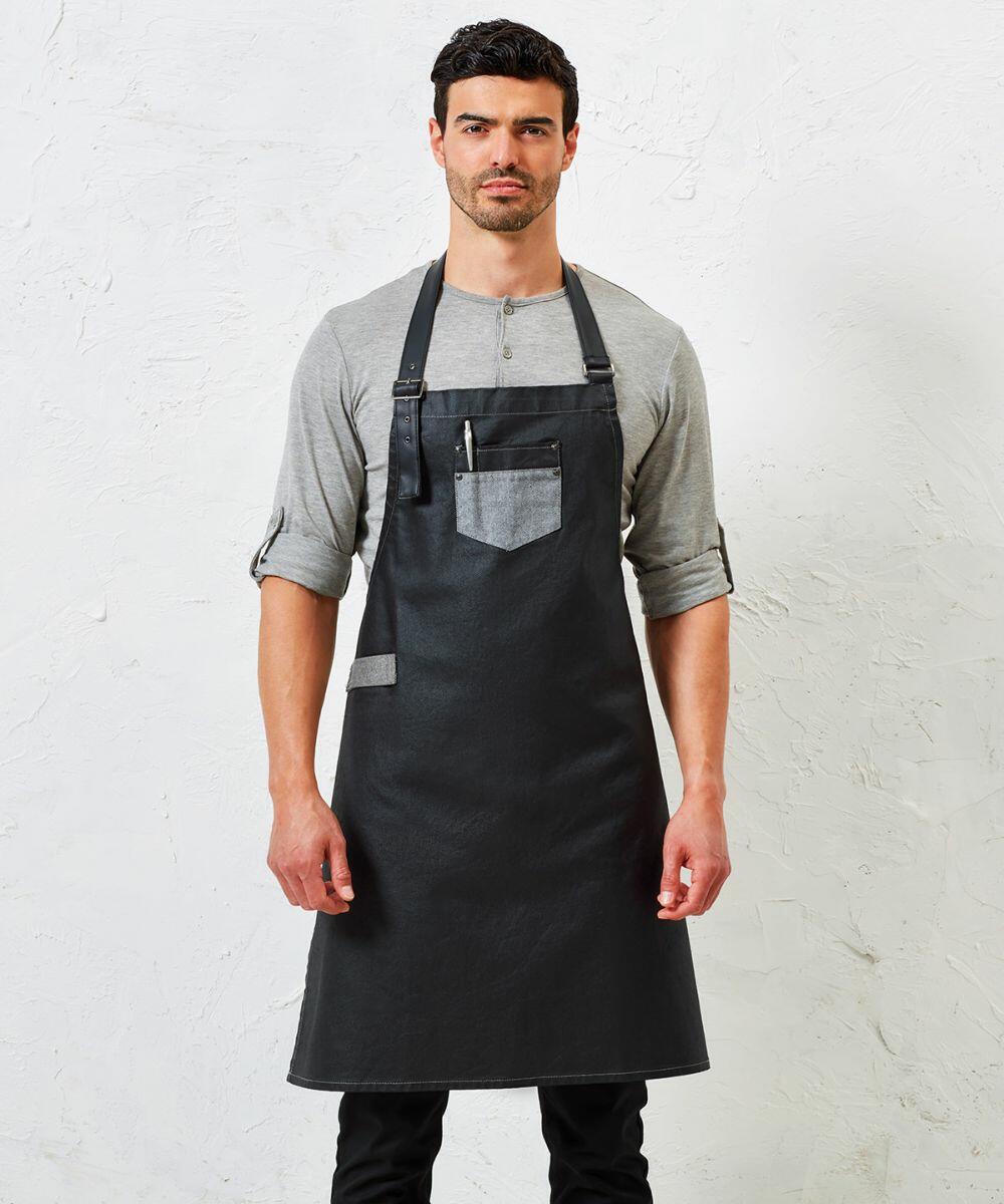 A man stands against a plain white background wearing a gray shirt and a black apron with a front pocket, which contains a pen. The apron is the PR136 by Premier.