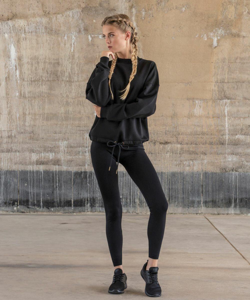 A woman stands in front of a textured, worn wall, wearing all-black athletic gear, including a Build Your Brand BY058 sweatshirt, leggings, and sneakers. She has long blonde hair styled in double braids. Her left hand is thoughtfully touching her chin as she looks to the side.