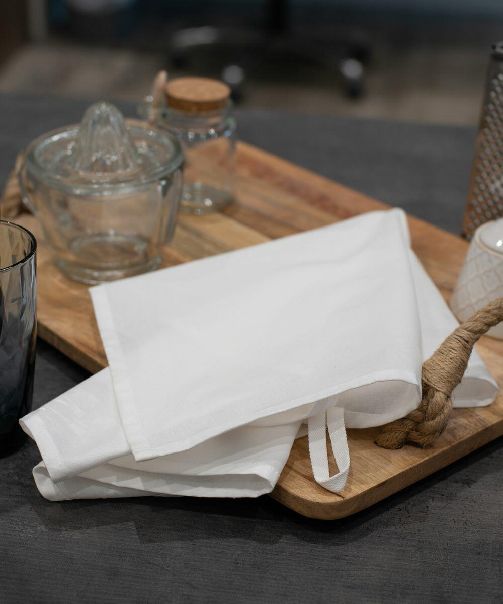 A folded white TC041 cloth napkin by Towel City rests on a wooden tray with a glass citrus juicer, small jars, and a decorative metal holder in the background.