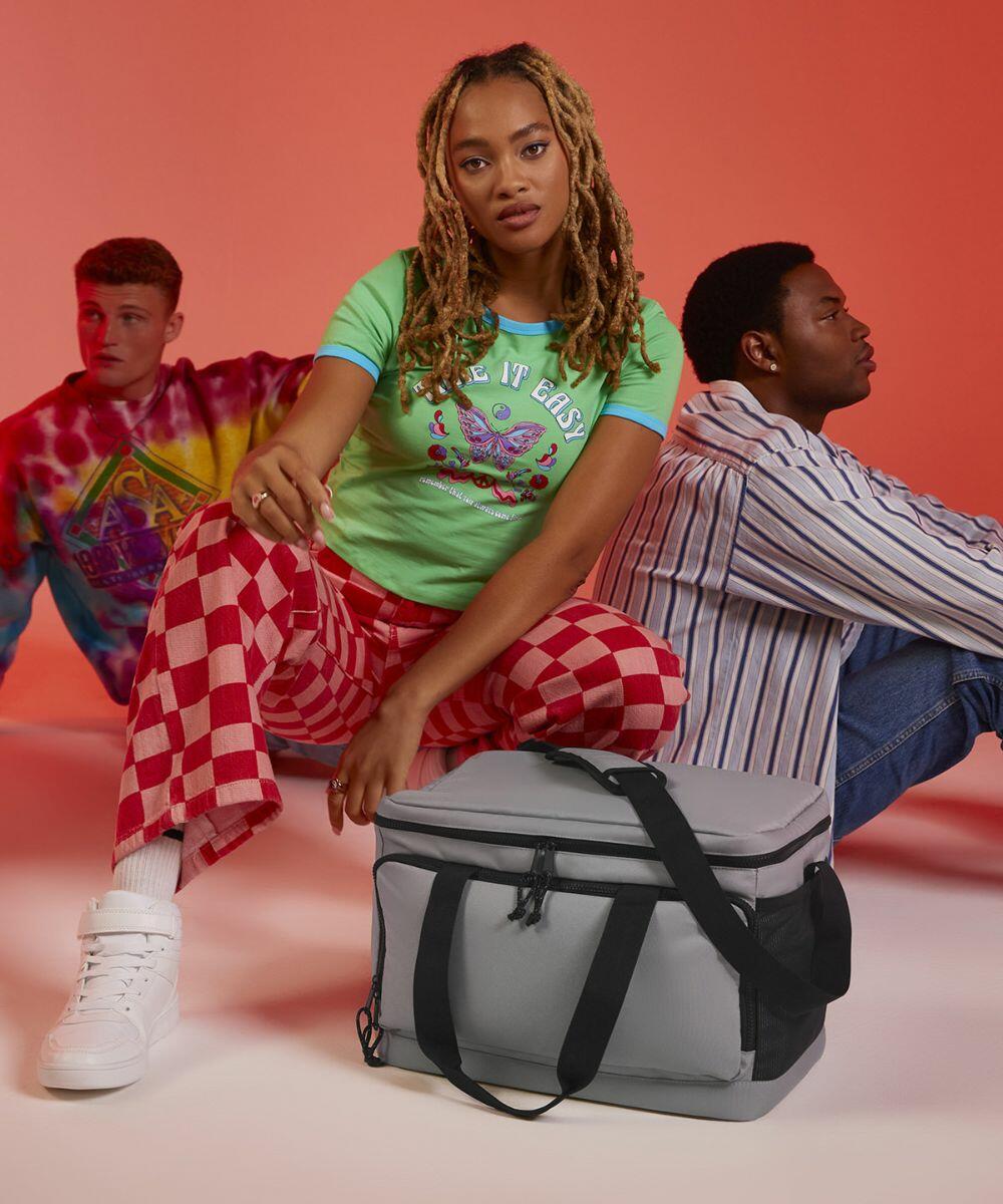 Three individuals posing against a gradient red background. The woman in front squats next to a large gray Bagbase BG290, wearing a green graphic tee and red checkered pants. A person in a seatbelt-pattern sweater and another in a striped shirt sit behind her.