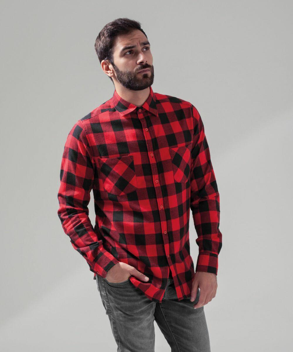 A man with short dark hair and a beard is wearing a red and black checkered BY031 flannel shirt by Build Your Brand and jeans. He is standing against a plain light gray background with his left hand in his pocket and looking slightly upward.