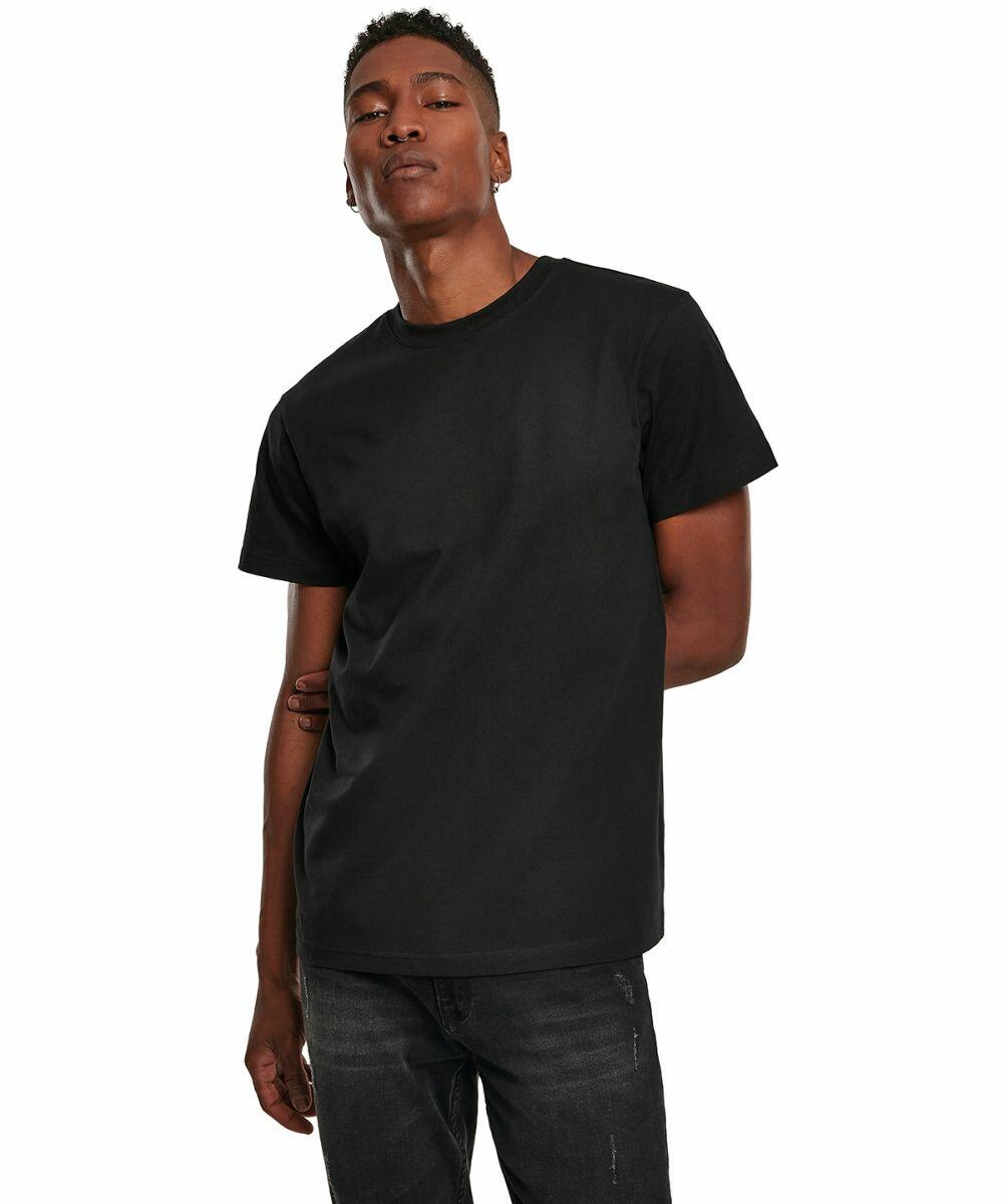A person stands facing forward with one hand resting on their other arm, wearing a plain black t-shirt and dark jeans from the Build Your Brand BY123 collection.