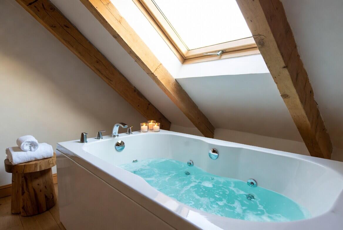 Modern straight double-ended white whirlpool spa bath with bubbling water in a bright upstairs attic bathroom with large skylight, wooden beams and candles