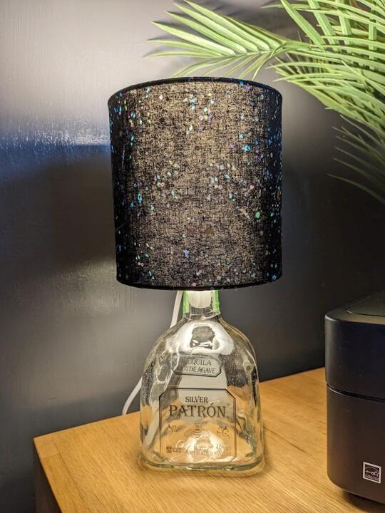 A handmade drinks bottle lamp with a clear tequila bottle base and a handmade lampshade with specks of colour on black fabric. The light is turned on.
