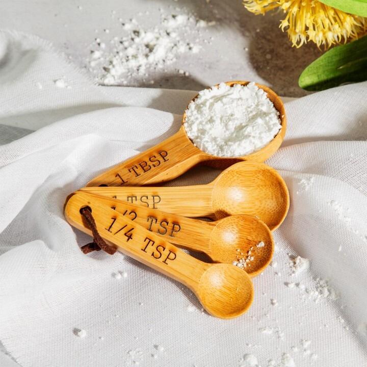 An image of a set of four bamboo measuring spoons laid out on a white surface with flour in the largest spoon