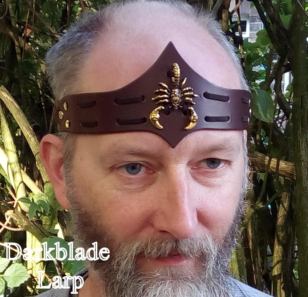 Face of a bearded man wearing a brown leather headband with a brass scorpion laced to it. The headband peaks to a point top and bottom on the centreline.