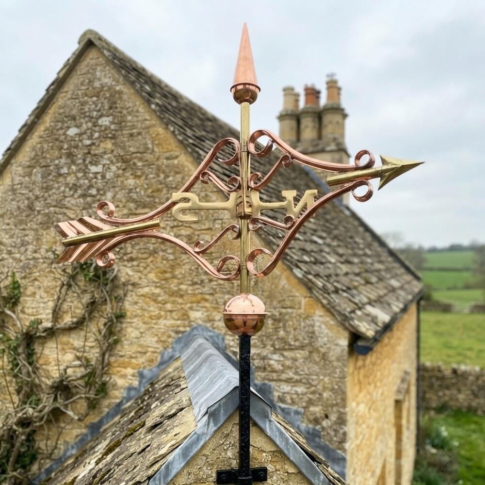 Medium copper Victorian arrow weathervane on a stone building showing scrolled copper detail and integrated cardinal points