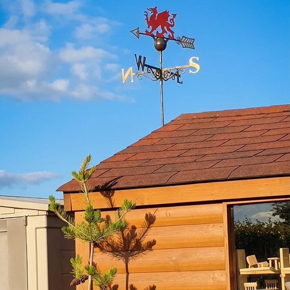 Welsh red dragon weathervane at ridge of cedar shingle shed roof with gold directional arms showing W E S, sunny garden setting