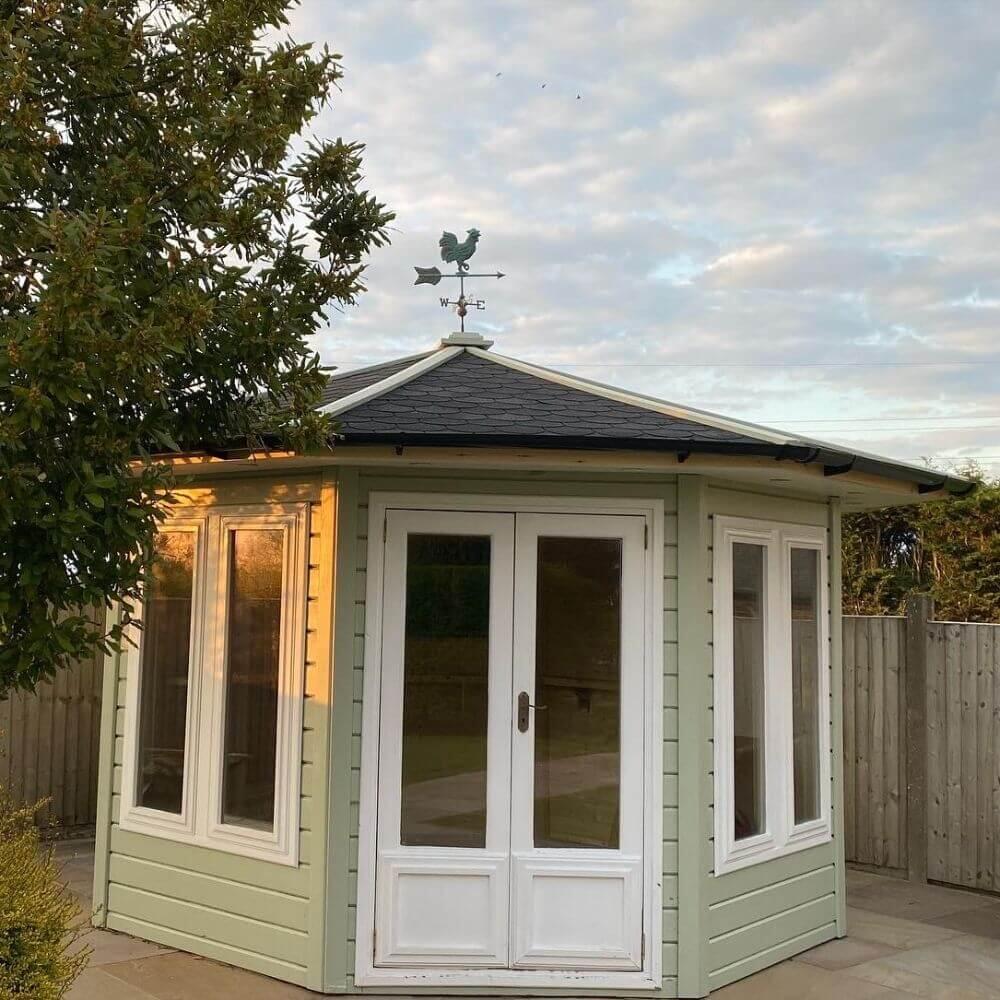 Front view of a pale green summerhouse with white double doors and the verdigris cockerel weathervane visible at the roof peak