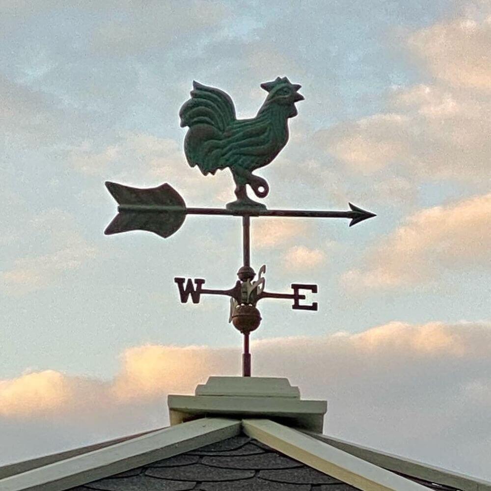 Close-up of aged copper verdigris cockerel weathervane on dark shingle roof with clouds behind