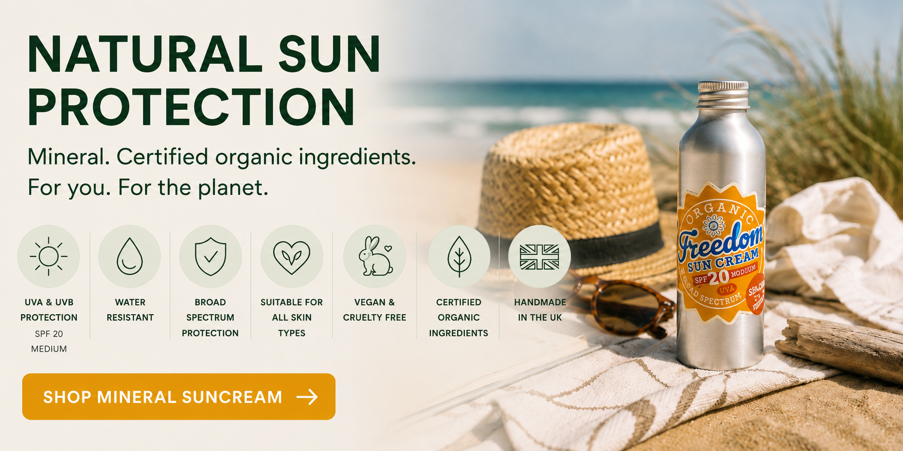 Banner promoting Freedom SPF20 Mineral Suncream on a beach with the headline "Natural Sun Protection". Features certified organic ingredients, broad-spectrum UVA and UVB protection, water resistance, vegan and cruelty-free formulation, and handmade in the UK. Bottle shown on a sandy beach beside a straw hat and sunglasses.