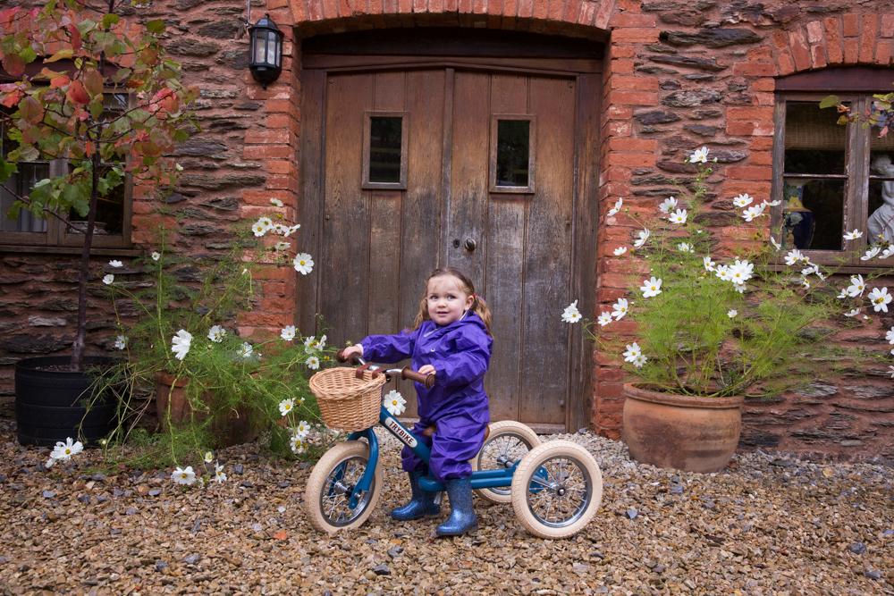 Child wearing a purple rainsuit on a green Trybike in front of a red brick house.