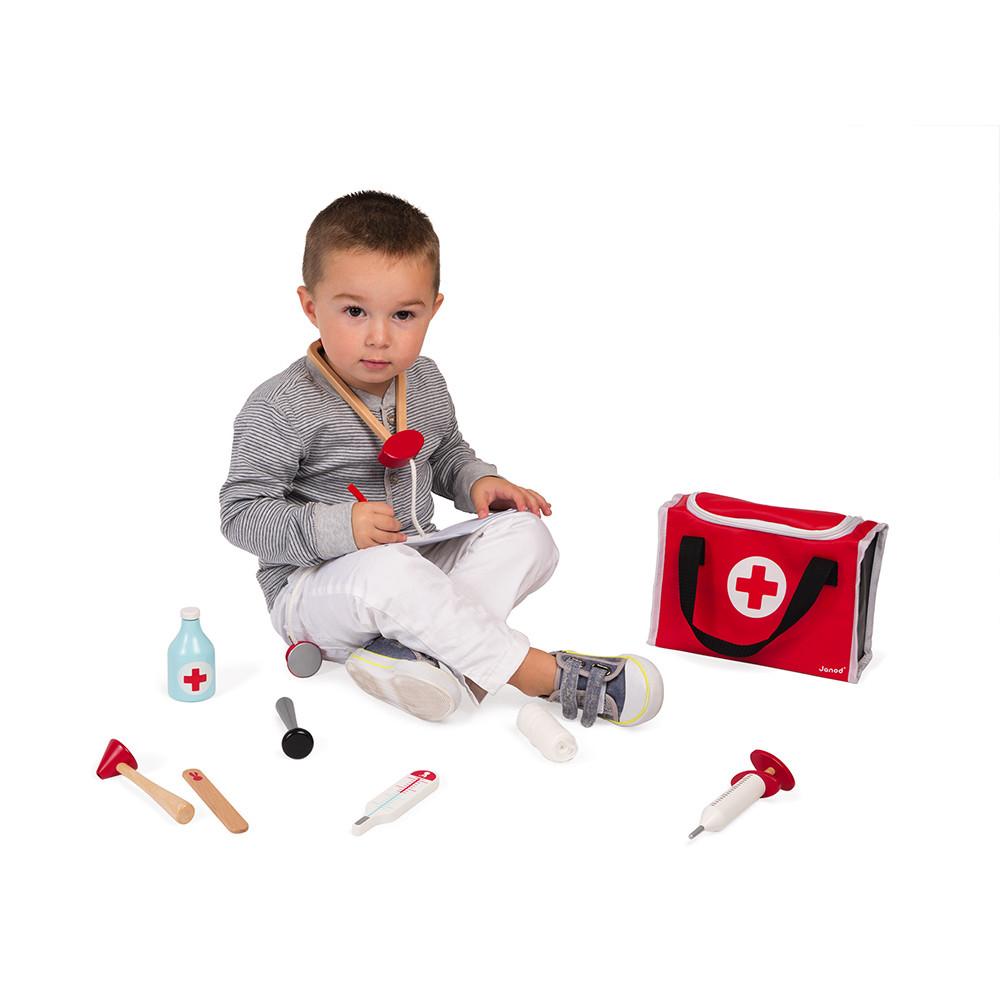 Young boy with wooden play stethoscope round his neck and surround by the other pieces of the doctor playset around him. White background.