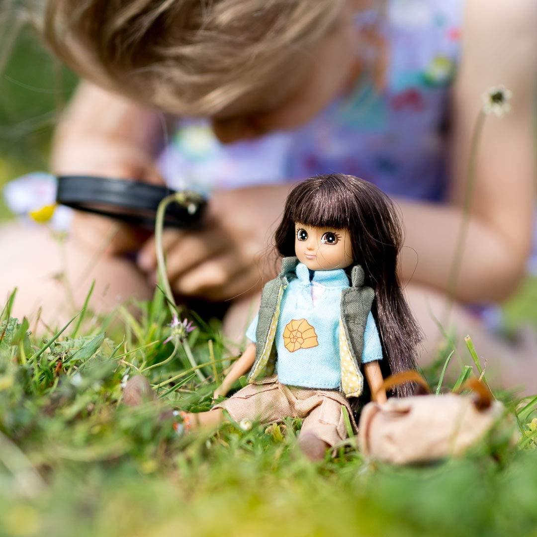 Dark-haired Lottie doll with a light blue t-shirt sitting on the grass with a young blonde-haired girl faded out in the background examining something closely with a magnifying glass..