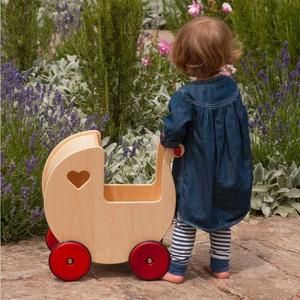 Back of a child in a dark blue denim dress beside a light wooden pram with red wheels. Flowers in the background.