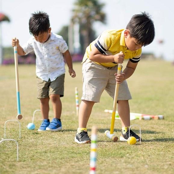 Two young boys with dark hair bent over playing croquest. The boy in the foreground has a yellow short-sleeved shirt and is hitting a yellow ball through the croquet hoop.