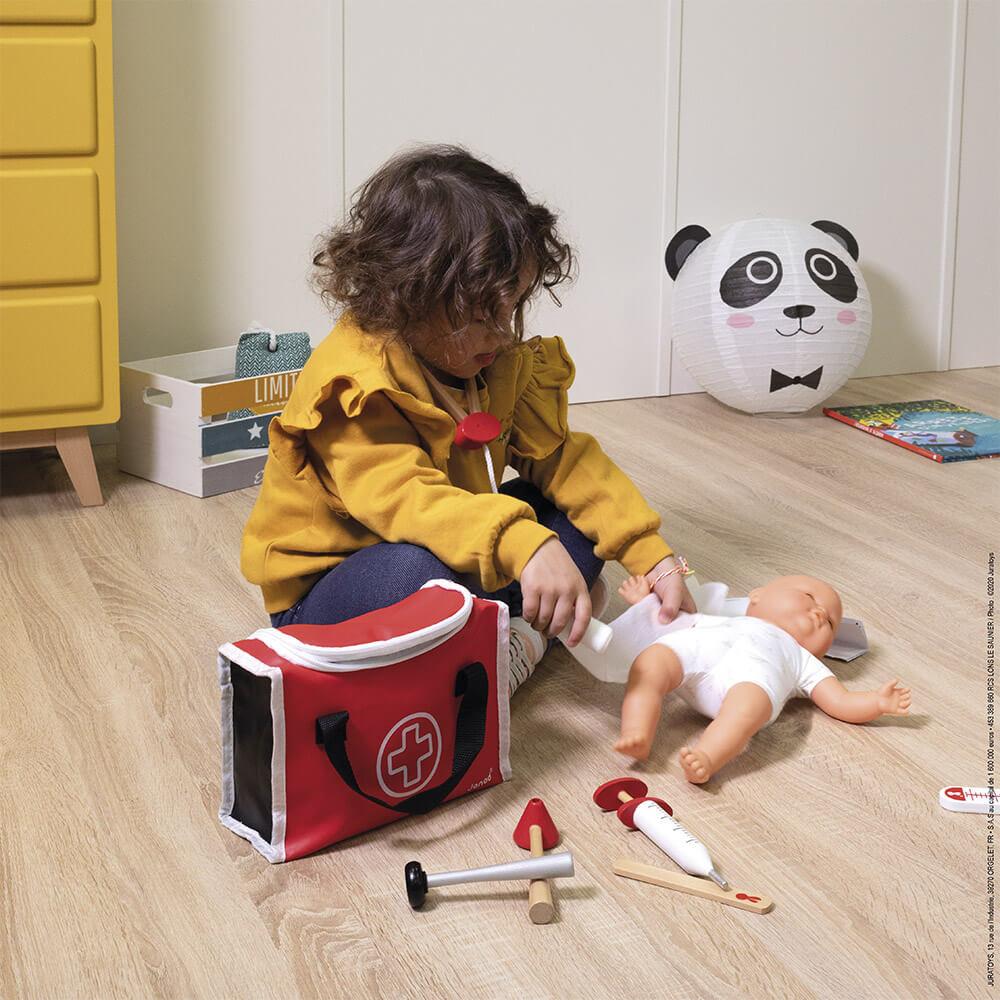 Child using the play doctor kit to attend to her dolly! Sitting on a wooden floor with the corner of a yellow chest of drawers in the background.