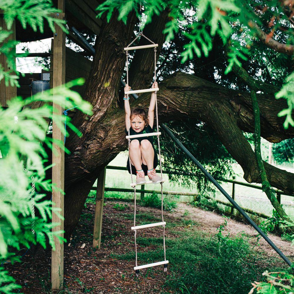 Girl with her arms which are above her head holding on to a wooden rope ladder with white ropes hanging from a thick-trunked tree.