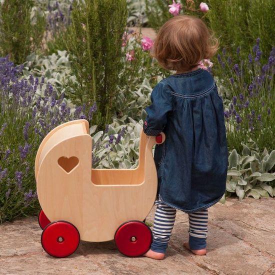 Young child standing with natural wood coloured pram with red wheels.