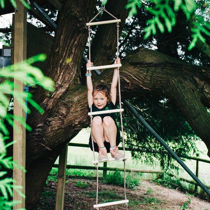 Girl on a wooden rope swing with pale wooden rungs and white rope attached to large tree. There is green foliage around and int he background. The girl has fair hair and she has her arms stretched out above her and her bare legs are bent.