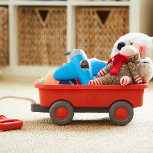 An orange coloured children's pull-along cart or wagon by Green Toys. The cart has grey wheels and a rope with an orange handle to pull it along. There is a cream toy bear and a blue toy aeroplane in the wagon. Cream carpet and hessian storage baskets in a unit behind.