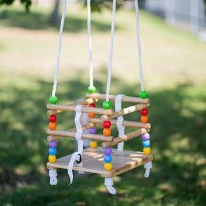 Wooden cradle swing with colourful wooden beads on the rope at each corner. The attached four white ropes go off the picture at the top. The background is a blurry shaded green garden.