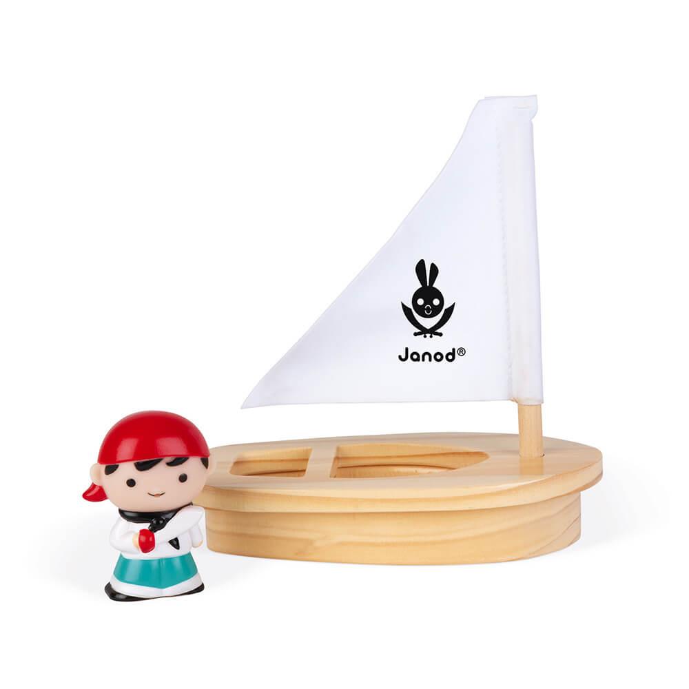 Wooden toy boat with white sail and the word 'Janod' written on the sail with a small pirate figure to the front left of the image. White background.